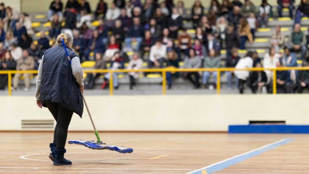 A person mopping a gymnasium floor with a blurred audience in the background
